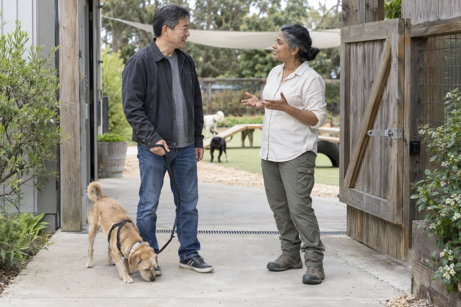 An owner and caregiver talking at drop-off while a tan dog sniffs beside them on a loose lead